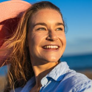 Happy woman holding pink sun hat and smiling at beach with ocean and blue sky in background during golden hour