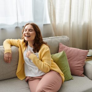 Middle aged woman sitting gracefully atop a sofa next to a large window.